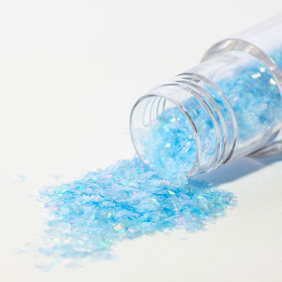 Transparent burk med blå iriserande glitterflakes som hälls ut på vit bakgrund.