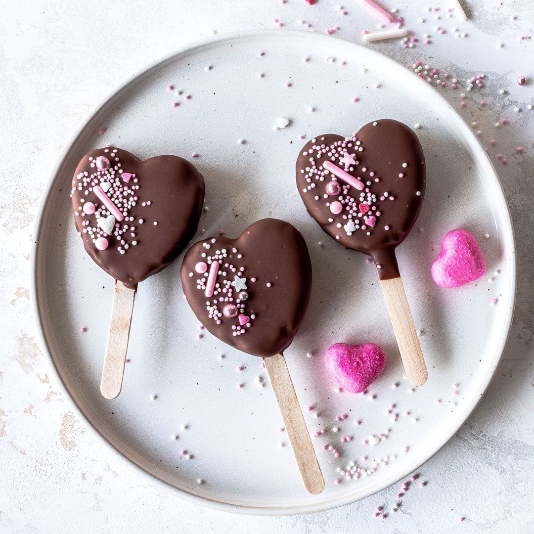 Chokladhjärtan på pinne dekorerade med rosa strössel på ett vitt fat.